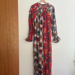 Puede incluir: Mono de manga larga con un diseño patchwork único. La prenda presenta una combinación de patrones de cuadros rojos, blancos y azules, junto con una sección de tela roja con gráficos de números coloridos. El mono tiene puños y tobillos con volantes.