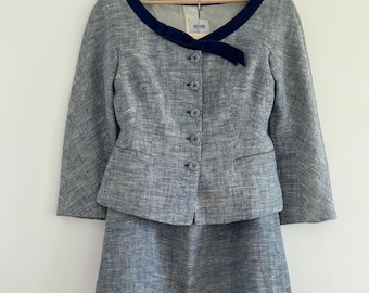 Puede incluir: Un traje de dos piezas azul claro con un tejido texturizado. La chaqueta tiene un ribete azul marino alrededor del cuello y un lazo a juego. La falda es de color azul claro a juego. El traje está colgado en una percha de madera.