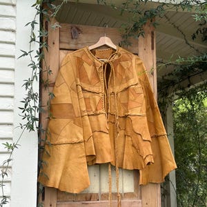 May include: A mustard yellow leather jacket featuring a patchwork design of various leather shapes. The jacket has long, flared sleeves, a round neckline, and a braided leather tie closure at the front. The construction includes visible seams.