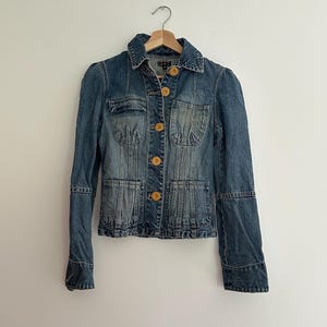 May include: A denim jacket with a classic design. The jacket features a collared neckline, button-down front, and multiple pockets. The jacket has a faded wash and light-colored buttons. The jacket is hanging on a wooden hanger.