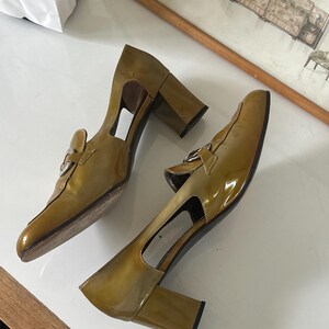 Puede incluir: Un par de zapatos de tac&oacute;n de charol verde oliva. Los zapatos tienen punta cuadrada, hebilla plateada y tac&oacute;n grueso. Los zapatos tienen un dise&ntilde;o recortado en los laterales.