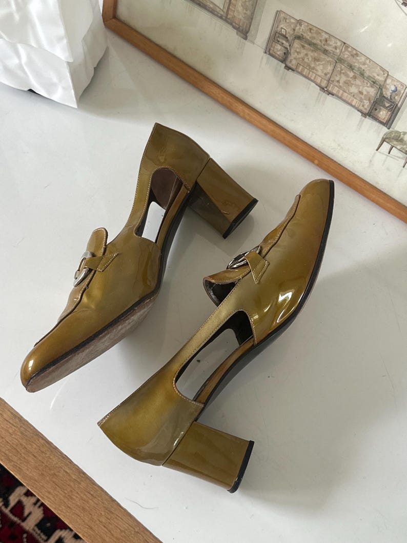 Puede incluir: Un par de zapatos de tac&oacute;n de charol verde oliva. Los zapatos tienen punta cuadrada, hebilla plateada y tac&oacute;n grueso. Los zapatos tienen un dise&ntilde;o recortado en los laterales.