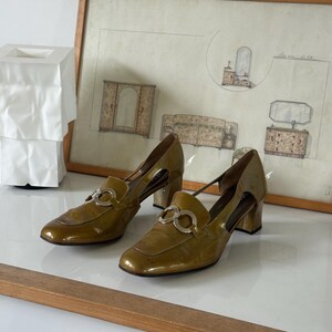 Puede incluir: Un par de zapatos de tac&oacute;n de charol verde oliva con herrajes plateados. Los zapatos tienen punta cuadrada y tac&oacute;n ancho. Se exhiben sobre una superficie blanca con un dibujo enmarcado al fondo.