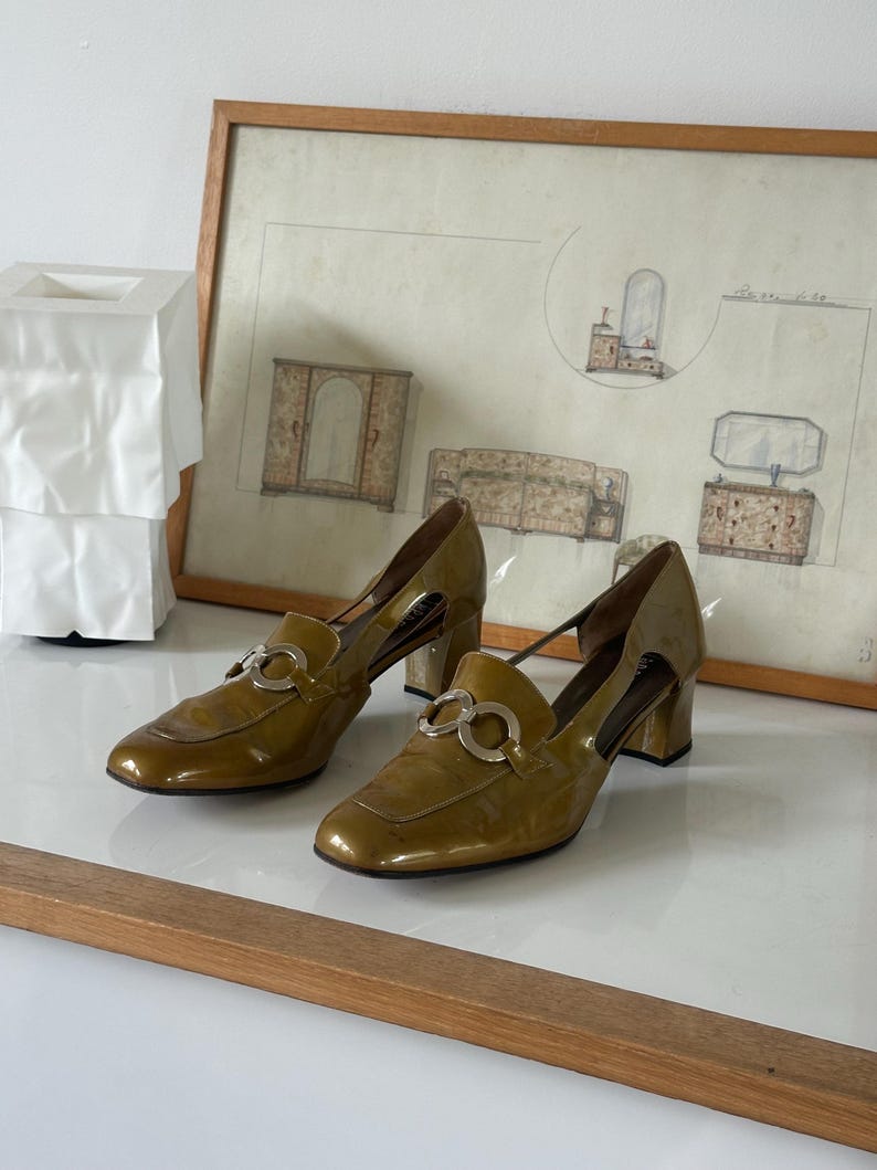 Puede incluir: Un par de zapatos de tac&oacute;n de charol verde oliva con herrajes plateados. Los zapatos tienen punta cuadrada y tac&oacute;n ancho. Se exhiben sobre una superficie blanca con un dibujo enmarcado al fondo.