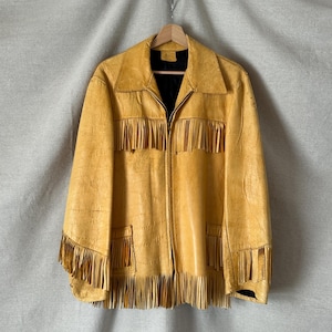 May include: A mustard yellow leather jacket featuring a full-length front zip, a pointed collar, and long fringe detailing across the chest, sleeves, and hem. The jacket includes two front pockets and a black interior lining, hanging on a wooden hanger.