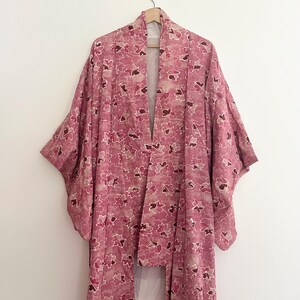 May include: A pink floral kimono hangs on a wooden hanger. The kimono features a repeating pattern of pink and burgundy flowers on a light pink background. The garment has wide sleeves and a long, flowing design.