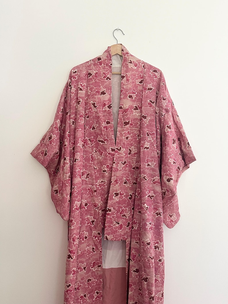 May include: A pink floral kimono hangs on a wooden hanger. The kimono features a repeating pattern of pink and burgundy flowers on a light pink background. The garment has wide sleeves and a long, flowing design.