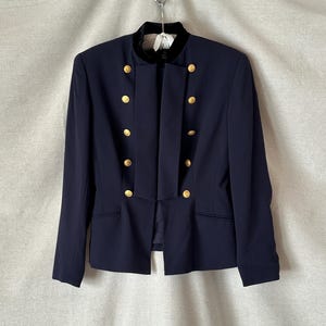 May include: A navy blue blazer featuring a mandarin collar with black velvet trim, long sleeves, and a double-breasted front closure with eight gold-toned buttons. The jacket has a structured silhouette with two vertical panels and two side pockets.