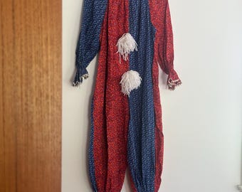 以下が含まれることがあります： 赤と青の花柄のヴィンテージピエロコスチューム。長袖のジャンプスーツは、片側に赤、もう片側に青の分割デザインになっています。白いポンポンとフリルの袖口が、遊び心のある衣装にディテールを加えています。