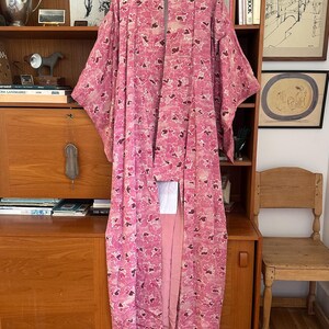 May include: A pink floral patterned kimono hangs on a wooden hanger. The kimono features a repeating pattern of pink and dark red flower-like shapes. The garment is long and flowing, with wide sleeves. The background includes a wooden cabinet and shelves.
