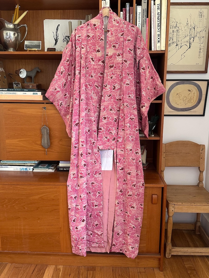 May include: A pink floral patterned kimono hangs on a wooden hanger. The kimono features a repeating pattern of pink and dark red flower-like shapes. The garment is long and flowing, with wide sleeves. The background includes a wooden cabinet and shelves.