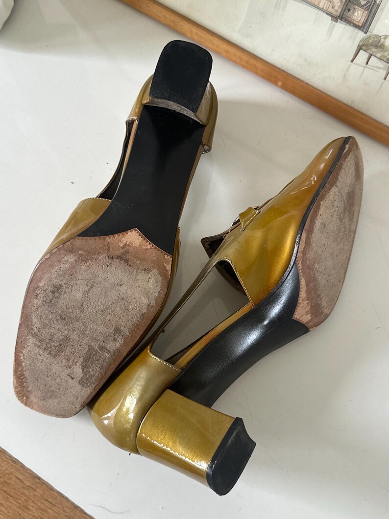 Puede incluir: Un par de zapatos vintage de charol dorado con punta cuadrada y tac&oacute;n ancho. Los zapatos tienen una correa en la parte superior del pie y una suela negra. Los tacones miden aproximadamente 7,6 cm de alto.
