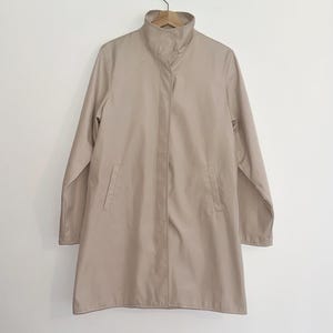 May include: A beige, long-sleeved coat with a high collar and two front pockets. The coat is hanging on a wooden hanger against a white background. The coat appears to be made of a lightweight fabric.