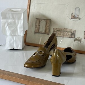 Puede incluir: Zapatos de tac&oacute;n de charol verde oliva con herrajes plateados. Los zapatos tienen punta afilada y tac&oacute;n cuadrado. Un dibujo enmarcado de muebles est&aacute; en el fondo.