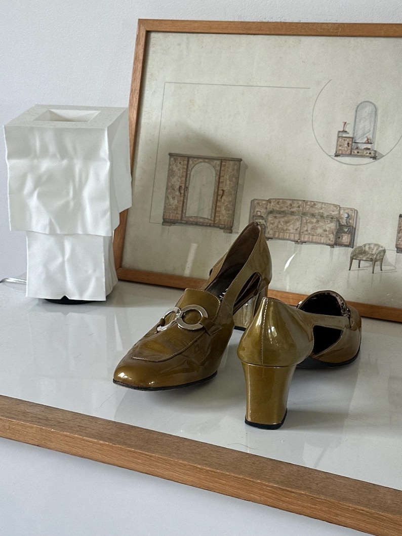 Puede incluir: Zapatos de tac&oacute;n de charol verde oliva con herrajes plateados. Los zapatos tienen punta afilada y tac&oacute;n cuadrado. Un dibujo enmarcado de muebles est&aacute; en el fondo.