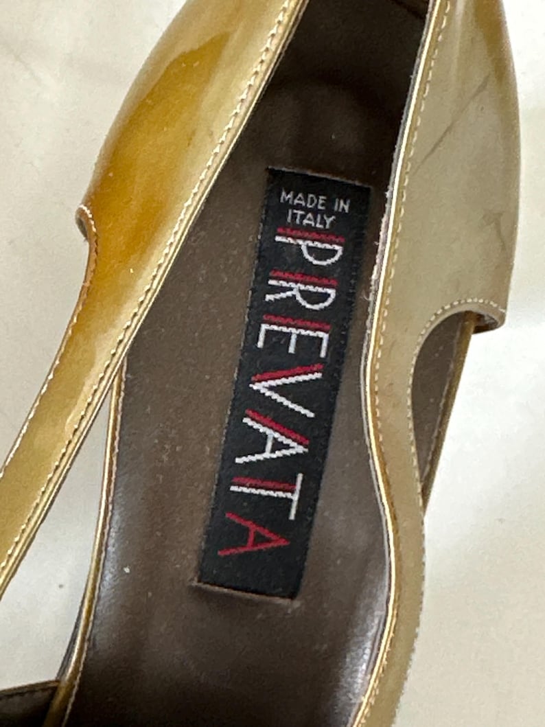 Puede incluir: Primer plano de un zapato de tac&oacute;n alto dorado. El zapato tiene un acabado brillante y un dise&ntilde;o recortado. El interior presenta una etiqueta que dice "MADE IN ITALY" y el nombre de la marca "PREVATA".