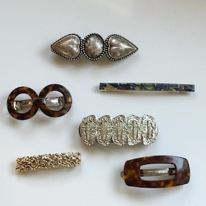 Könnte beinhalten: Eine Sammlung von Haarspangen in verschiedenen Stilen. Enthält eine silberfarbene Spange mit herzförmigen Akzenten, eine Spange mit Schildpattmuster und eine goldfarbene Blumen-Spange. Weitere Spangen weisen silberne und gemusterte Designs auf.