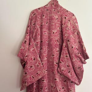 May include: A pink kimono with a floral pattern hangs from a metal hanger. The kimono features a repeating design of stylized flowers in shades of pink and burgundy. The garment has wide sleeves and a high collar.