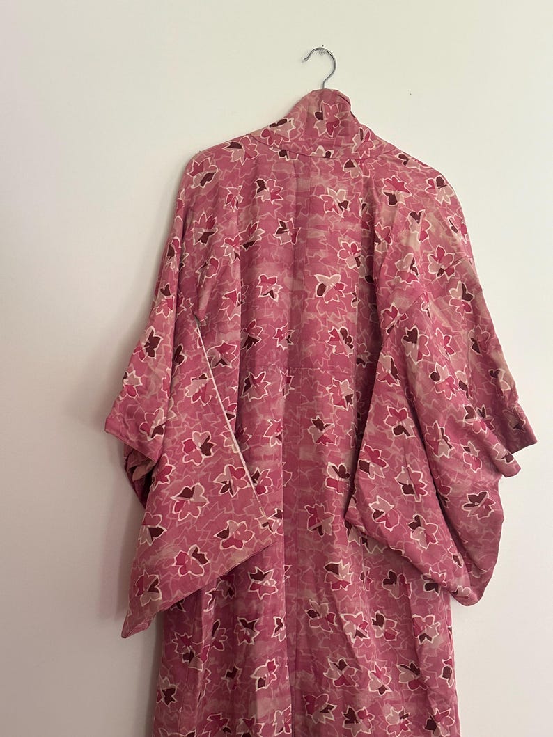 May include: A pink kimono with a floral pattern hangs from a metal hanger. The kimono features a repeating design of stylized flowers in shades of pink and burgundy. The garment has wide sleeves and a high collar.