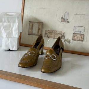 Puede incluir: Un par de mocasines de tac&oacute;n de cuero verde oliva con detalles plateados. Los zapatos tienen punta cuadrada y tac&oacute;n bajo. La palabra "PREVATE" es visible en el lateral. Un dibujo arquitect&oacute;nico enmarcado en el fondo.