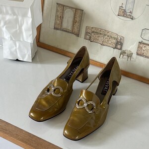 Puede incluir: Un par de zapatos de charol verde oliva con herrajes plateados. Los zapatos tienen punta cuadrada y tac&oacute;n de bloque peque&ntilde;o. Los zapatos se exhiben sobre una superficie blanca.