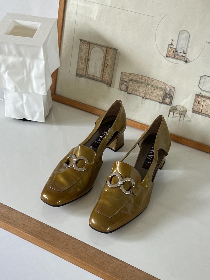 Puede incluir: Un par de zapatos de charol verde oliva con herrajes plateados. Los zapatos tienen punta cuadrada y tac&oacute;n de bloque peque&ntilde;o. Los zapatos se exhiben sobre una superficie blanca.