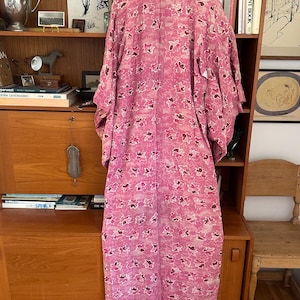 May include: A pink floral patterned kimono hangs on a wooden shelf. The kimono features a repeating pattern of pink and white flowers with brown accents. The garment is made of a lightweight fabric and has wide sleeves.