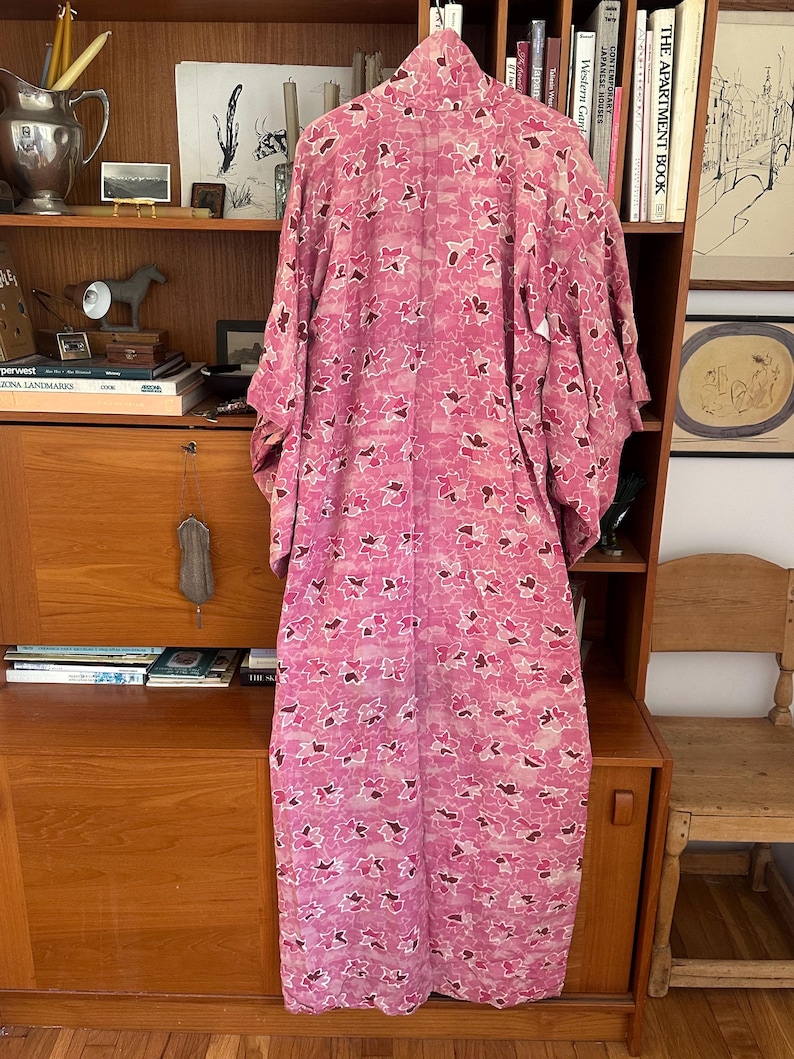 May include: A pink floral patterned kimono hangs on a wooden shelf. The kimono features a repeating pattern of pink and white flowers with brown accents. The garment is made of a lightweight fabric and has wide sleeves.