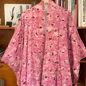 May include: A pink floral patterned kimono hanging on a wooden hanger. The kimono features a repeating pattern of pink and burgundy flower shapes on a light pink background. The garment is displayed in front of a wooden bookshelf.