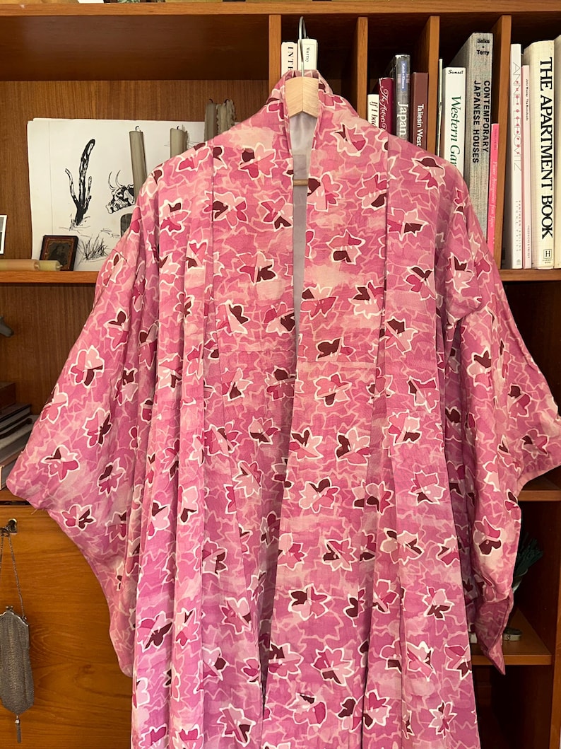 May include: A pink floral patterned kimono hanging on a wooden hanger. The kimono features a repeating pattern of pink and burgundy flower shapes on a light pink background. The garment is displayed in front of a wooden bookshelf.