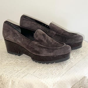 May include: A pair of dark brown suede platform loafers. The shoes have a rounded toe, a glossy dark brown platform sole, and a stacked wedge heel. The shoes are displayed on a white lace tablecloth.