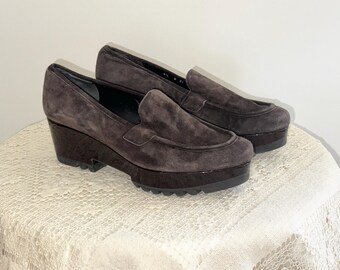 May include: A pair of dark brown suede platform loafers. The shoes have a rounded toe, a glossy dark brown platform sole, and a stacked wedge heel. The shoes are displayed on a white lace tablecloth.
