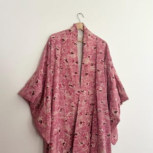 May include: A pink floral kimono hangs on a wooden hanger against a white wall. The kimono features a repeating pattern of pink and brown flower shapes. The garment has wide sleeves and a long, open front.