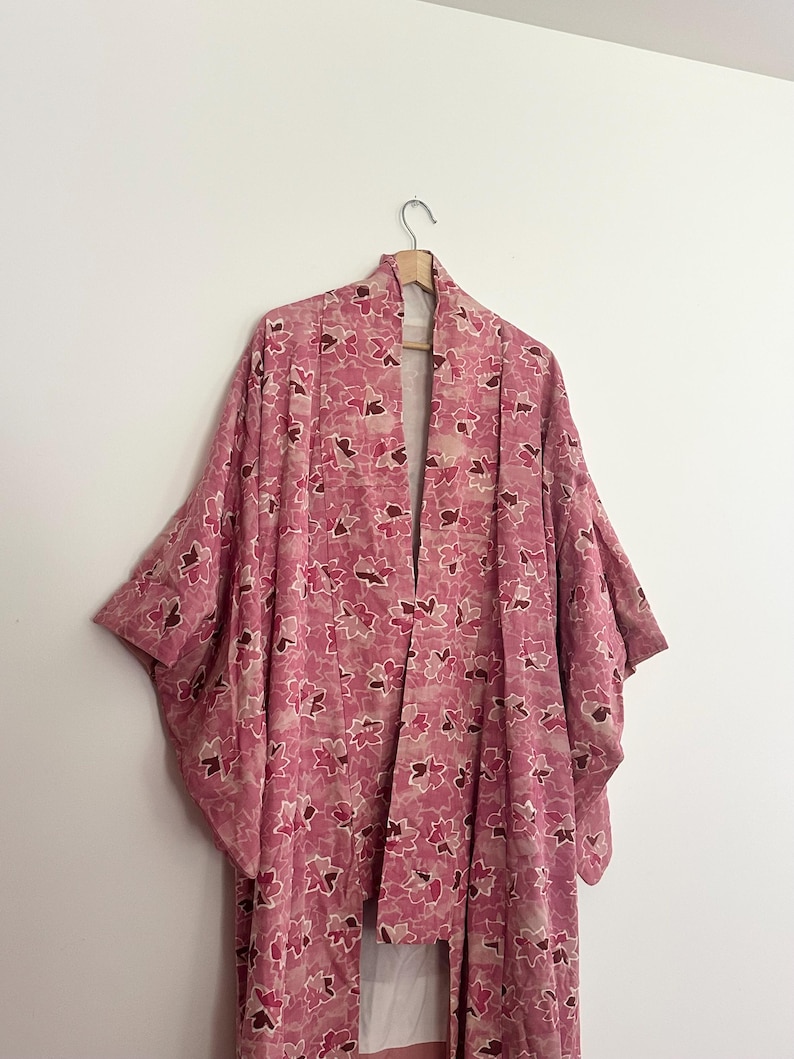May include: A pink floral kimono hangs on a wooden hanger against a white wall. The kimono features a repeating pattern of pink and brown flower shapes. The garment has wide sleeves and a long, open front.