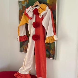 Puede incluir: Un colorido disfraz de payaso con una combinación de colores blanco, rojo y amarillo. El disfraz presenta un cuello con volantes, pompones rojos y puños con volantes. El atuendo está colgado en una percha de madera contra una pared con una pintura.