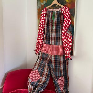 Puede incluir: Un colorido disfraz de payaso con mangas de lunares rojos y un corpiño y pantalón a cuadros. El disfraz tiene una cintura de cuadros rojos y blancos y parches de cuadros rojos en las rodillas. El disfraz está colgado en una percha de madera.