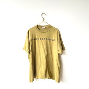 May include: A mustard yellow t-shirt with a row of black and white circles with the number "50" inside each circle.