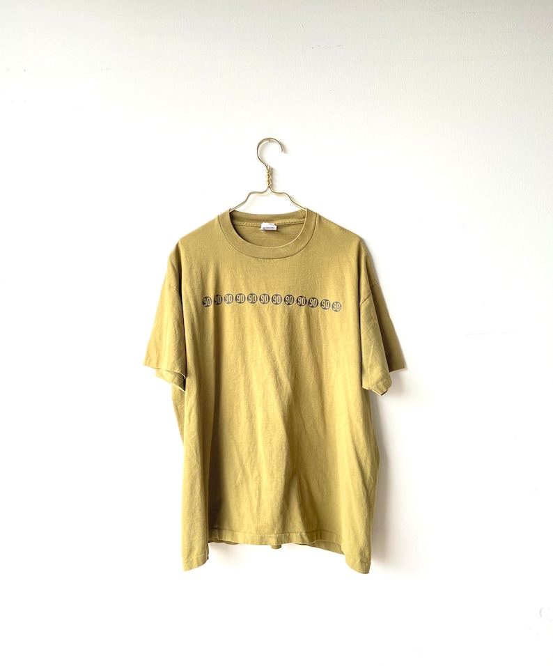 May include: A mustard yellow t-shirt with a row of black and white circles with the number "50" inside each circle.