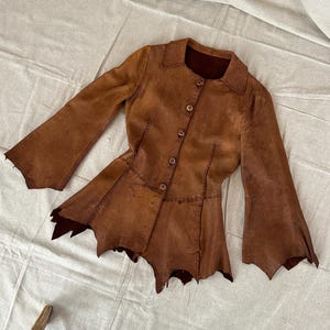 May include: A brown leather jacket featuring a pointed collar, long flared sleeves, and a button-down front. The garment has a fitted waist and a hemline with jagged, uneven edges. Visible stitching details run along the seams of the bodice.