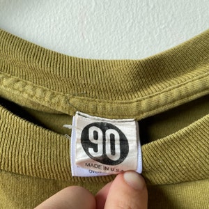May include: A green t-shirt with a white tag that says "90" in a circle and "Made in U.S.A."