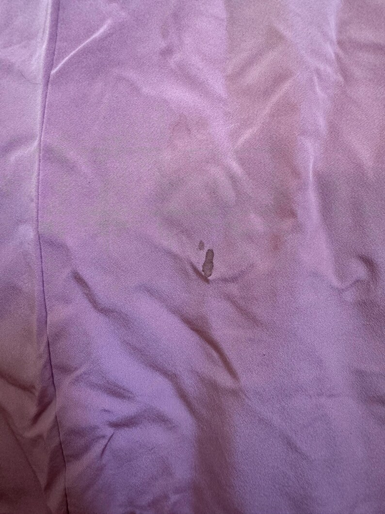 May include: Close-up of a lavender fabric with visible wrinkles and dark spots. The fabric appears to be a soft material, possibly a garment or textile. The image highlights the texture and color variations.