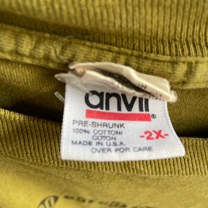 May include: A white tag with black text that reads "anvil" in a bold font. The tag also says "Pre-shrunk 100% Cotton/Coton Made in U.S.A. -2X- Over for care".