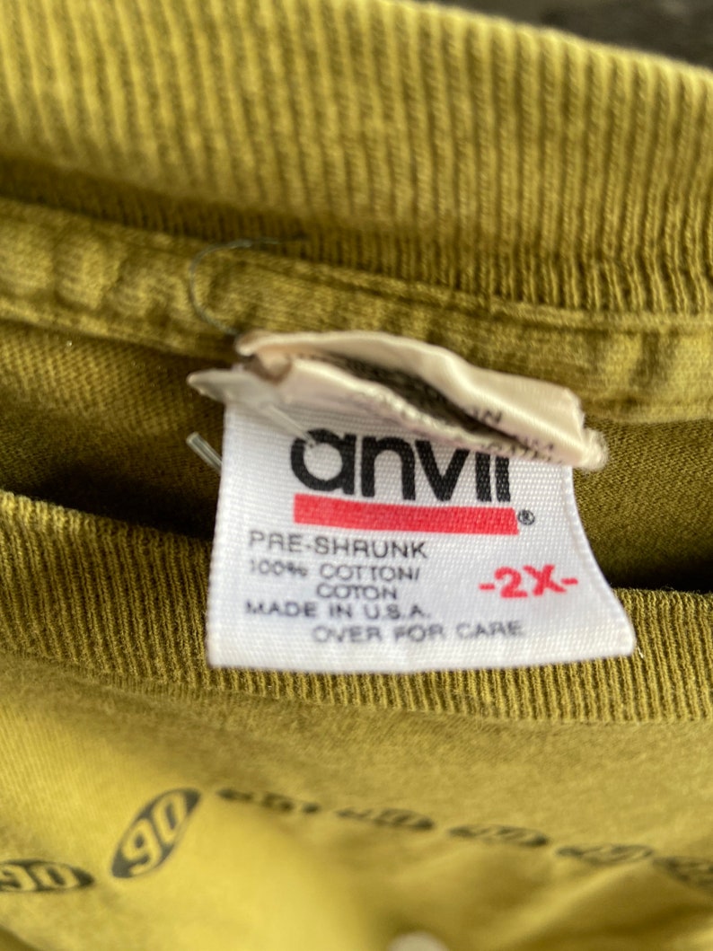May include: A white tag with black text that reads "anvil" in a bold font. The tag also says "Pre-shrunk 100% Cotton/Coton Made in U.S.A. -2X- Over for care".