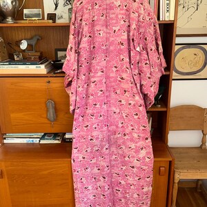 May include: A long, pink kimono with a floral pattern in shades of pink and brown hangs in front of a wooden cabinet. The kimono is made of a lightweight fabric and has wide sleeves. The background includes a wooden cabinet with books and decorative items.