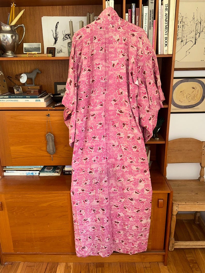 May include: A long, pink kimono with a floral pattern in shades of pink and brown hangs in front of a wooden cabinet. The kimono is made of a lightweight fabric and has wide sleeves. The background includes a wooden cabinet with books and decorative items.
