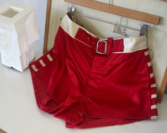 May include: High-waisted red satin shorts featuring a wide cream-colored waistband with a silver-toned metal buckle belt. The shorts include cream-colored vertical striped side panels and are displayed on a wooden hanger against a light background.