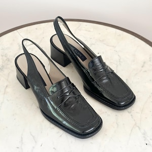 Puede incluir: Un par de mocasines slingback de cuero negro con punta cuadrada y tacón de bloque. Los zapatos tienen un acabado brillante y una correa en la parte superior del pie. La marca "ANNE KLEIN II" es visible en la plantilla.
