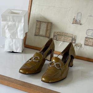 Puede incluir: Un par de mocasines de tac&oacute;n de charol verde oliva con detalles plateados. Los zapatos tienen punta cuadrada y cortes laterales. Los tacones miden aproximadamente 6,35 cm de alto.