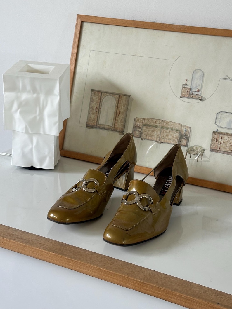 Puede incluir: Un par de mocasines de tac&oacute;n de charol verde oliva con detalles plateados. Los zapatos tienen punta cuadrada y cortes laterales. Los tacones miden aproximadamente 6,35 cm de alto.