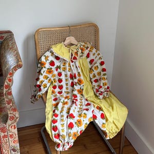 Puede incluir: Conjunto vintage de bebé compuesto por un pelele y una chaqueta a juego. El pelele presenta un fondo blanco con un estampado de fresas rojas y flores amarillas. La chaqueta y el pelele tienen detalles amarillos. El conjunto se exhibe en una silla de mimbre.